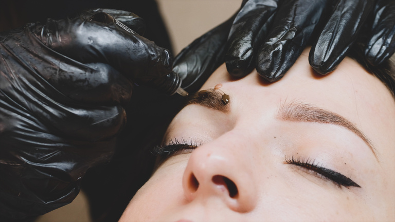 The master is adding some drops of the brown pigment on the client's eyebrow.